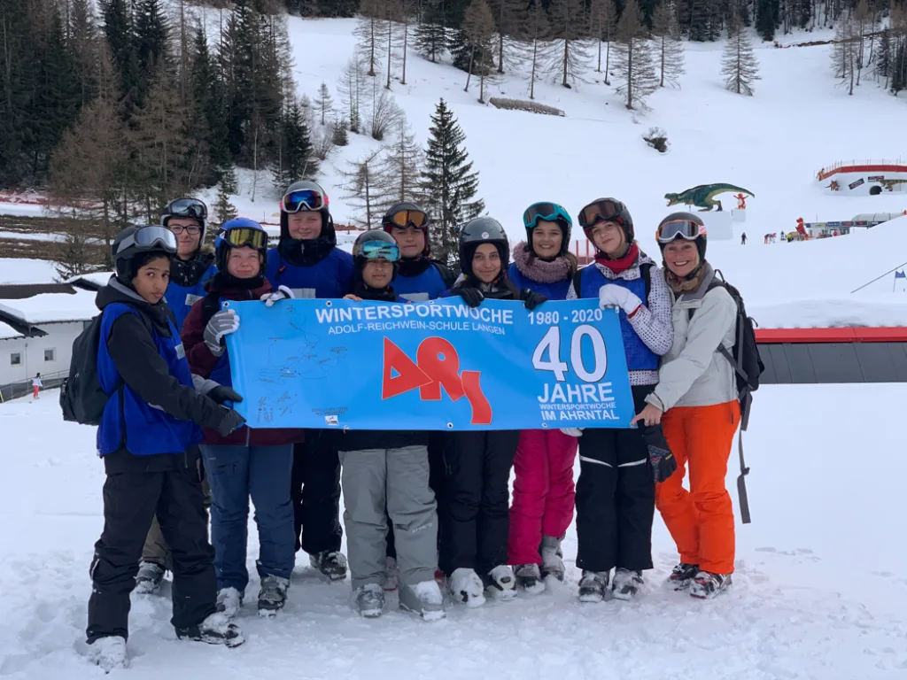 Die Gruppe hält das Banner, dahinter der Skihang mit Liftanlage.